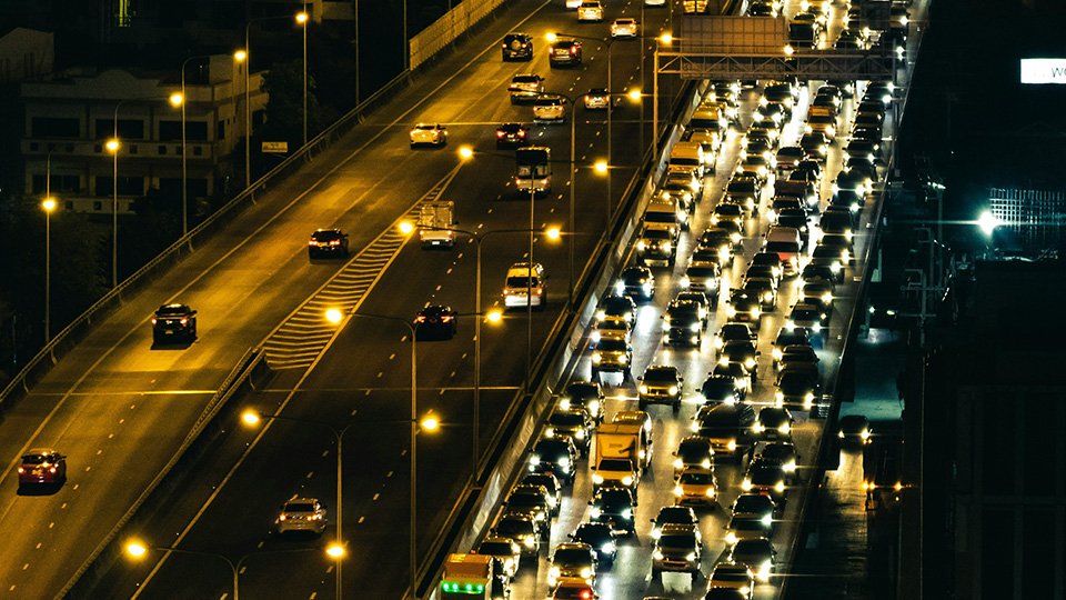 A Imagem mostra vias iluminadas, cheias de veículos na parte da noite.