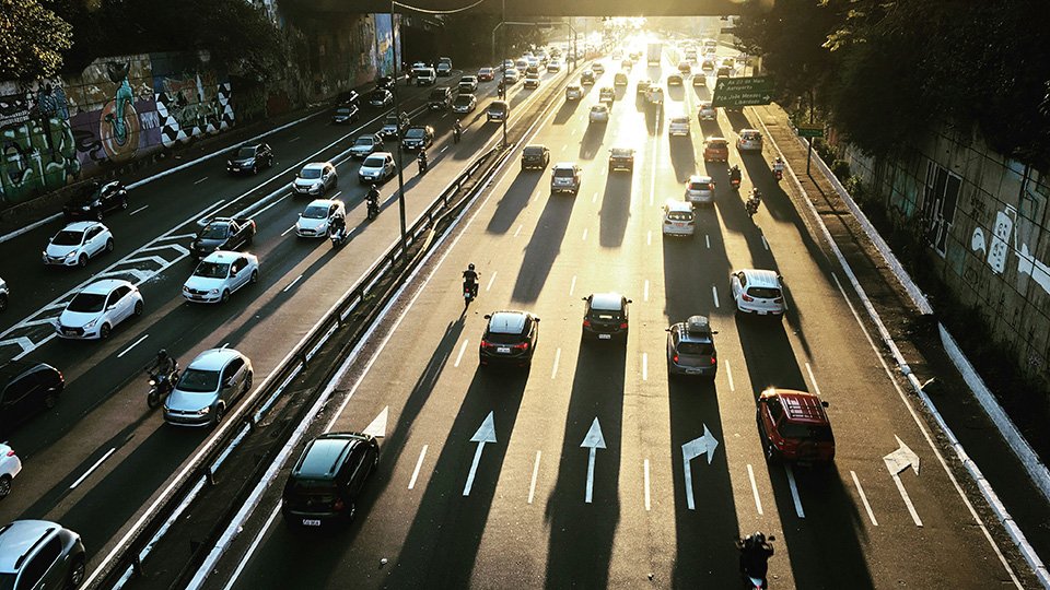 A imagem mostra uma via urbana larga com diversos carros e motos, dirigndo em direção ao horizonte