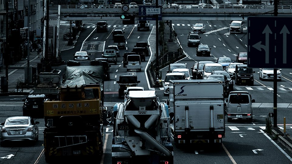 A Imagem tem uma via com tráfego intenso, em cores de tons mais frios. Com carros e caminhões.