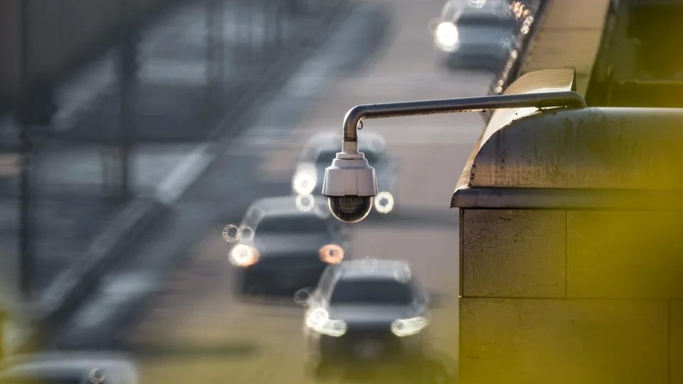 A Imagem mostra carros passando numa via, em desfoque, com o foco numa câmera que capta imagens dos veículos que passam
