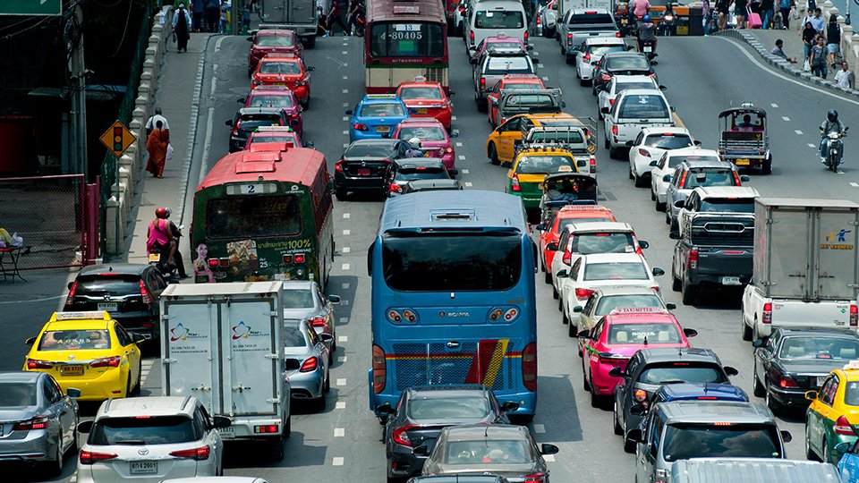 Uma via com trânsito intenso de veículos, no centro urbano com alguns outdoors e placas