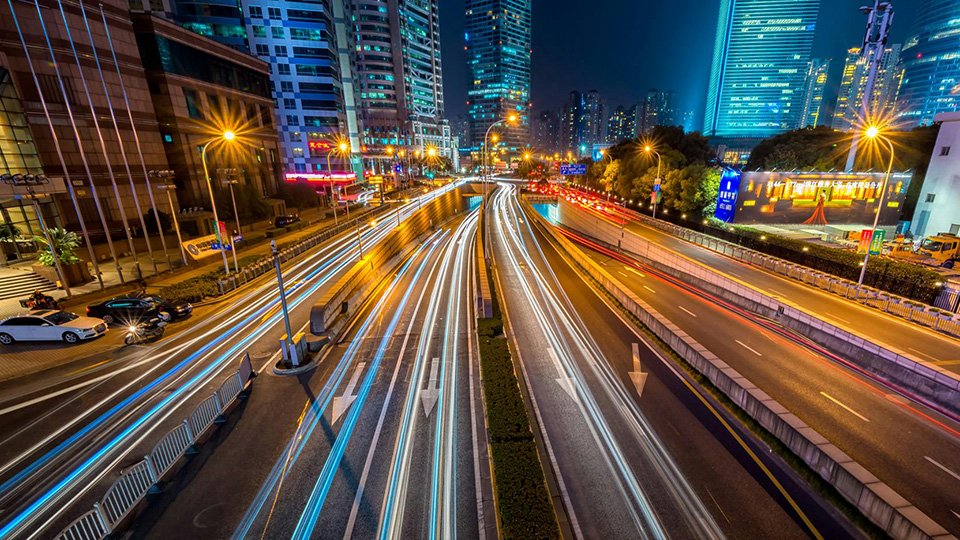 Imagem de uma cidade, à noite, com diversas luzes, e carros passando em alta velocidade pelas vias