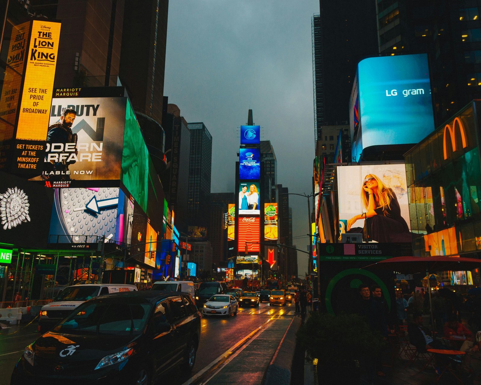 Uma rua, no fim de tarde, com vários outdoors e painéis digitais com propagandas, no meio da cidade, enquanto passam carros e outros veículos