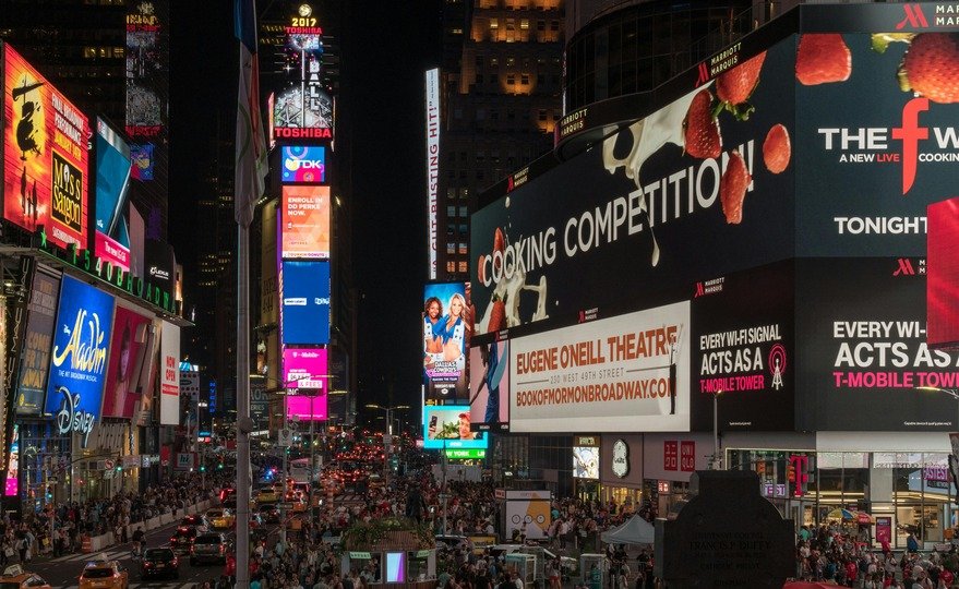 Rua comercial iluminada com fluxo de pedrestres e veículos, com diverso anúncios e paineis digitais