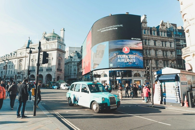 A imagem mostra uma rua, com pedrestres e veículos, próximos à edifícios e um grande painel digital com anúncios