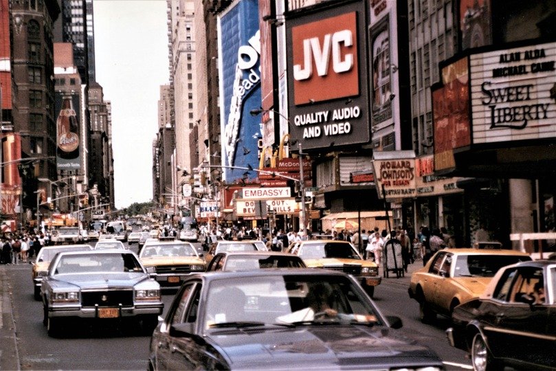 Veículos passando numa rua com diversas propagandas, outdoors e painéis de anúncios