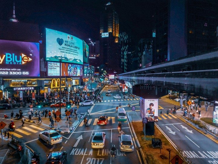 A imagem mostra um centro urbano com carros e pedrestres passando, lojas de rua, e propagandas como outdoors, e painéis digitais