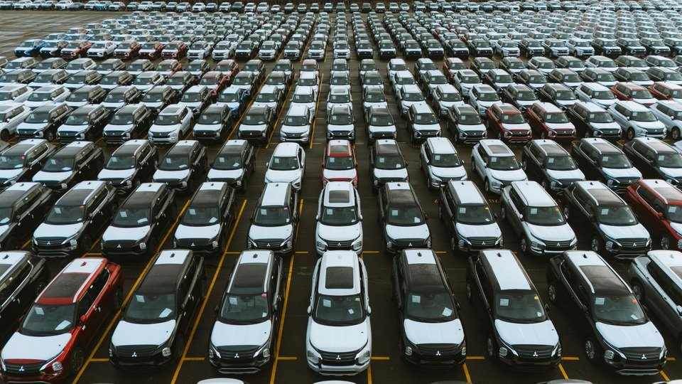 A imagem mostra diversos tipos de carros estacionados em fileiras , como em um estacionamento cheio, tendo os primeiros carros a cor branca/prata