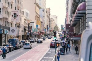 A imagem mostra uma rua com hotéis e edifícios com veículos e pedestres circulando, numa manhã