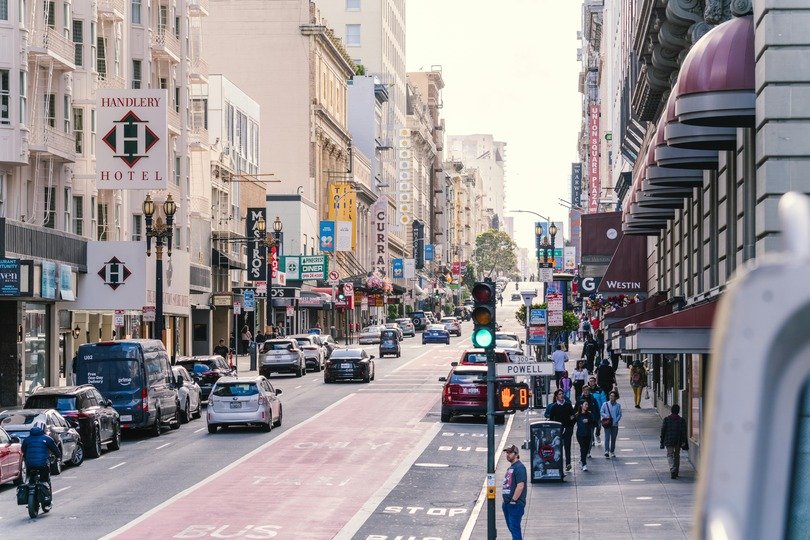 A imagem mostra uma rua com hotéis e edifícios com veículos e pedestres circulando, numa manhã