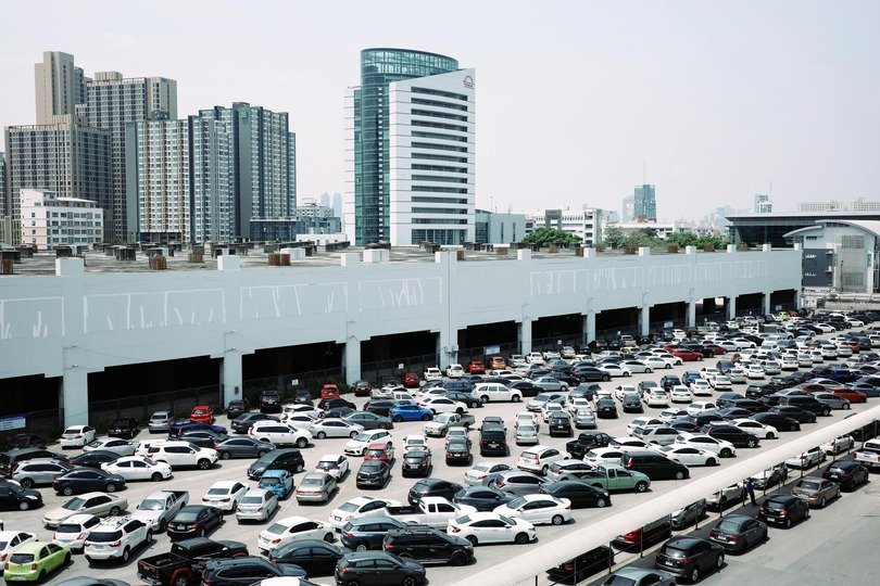 A Imagem mostra alguns edifícios, e um grande estacionamento cheio de carros parados de forma desorganizada no que parece ser um espaço grande de conferências ou reunião de pessoas